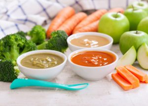 Three bowls of baby food placed around fresh vegetables and fruits