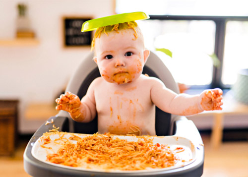 A baby on a high chair with food spilt all around and the bowl on the head.