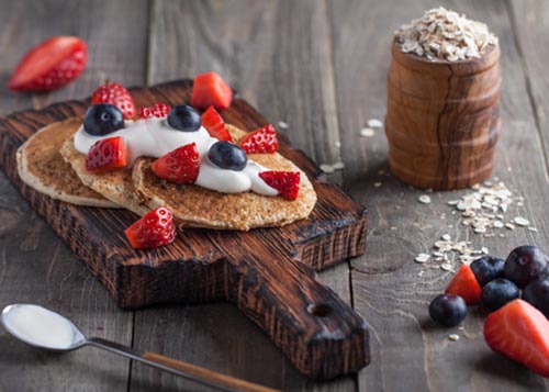 Pancakes with berries and yoghurt on top of a serving board with berries and a bowl of oats next to it.