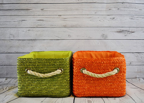 A green and orange storage basket made of wicker, lined with cushion, kept against the wall next to each other.
