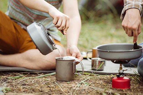 A person pouring water into a mug and another person cooking on a camp stove.