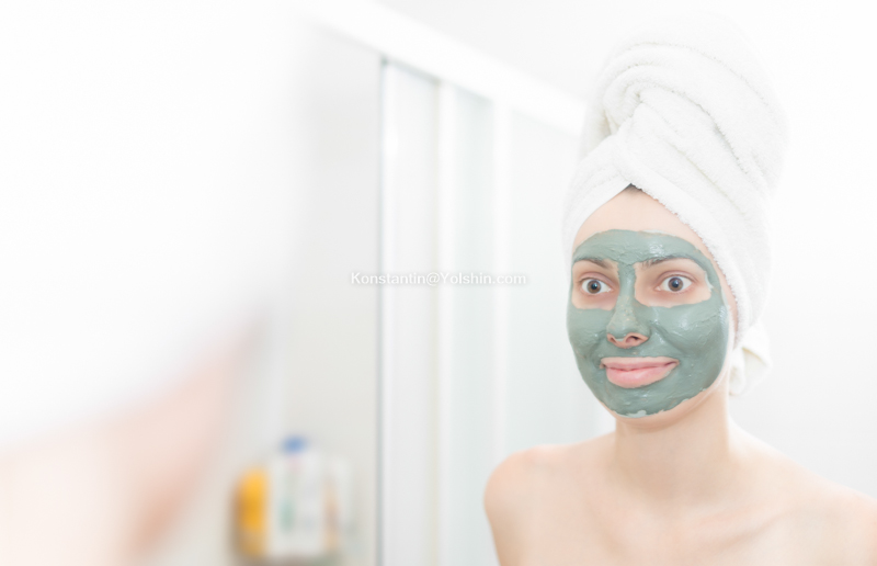 Woman with a clay mask on her face looking into the mirror 