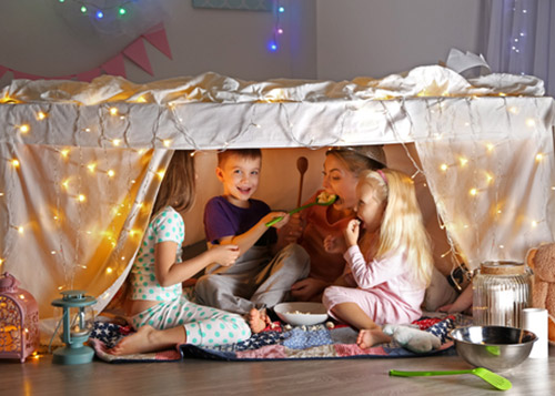 Mother and kids eating and having fun inside an indoor fort made from tables and sheets