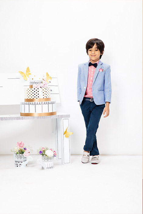 A boy wearing a pink shirt, checked blue blazer, navy blue pants, and white shoes standing next to a table with a cake