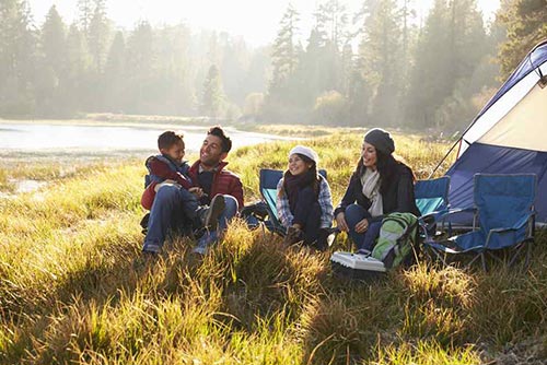 Family sitting on the grass outside their camping tent next to a stream