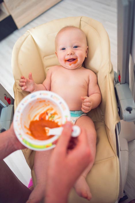 An adult feeding orange coloured food to a baby in a chair