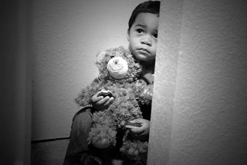 A scared child sitting in the corner, holding a teddy bear.