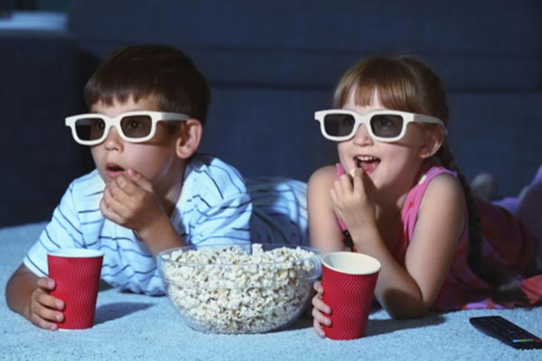 Two kids lying on a rug with popcorn and beverages watching a movie