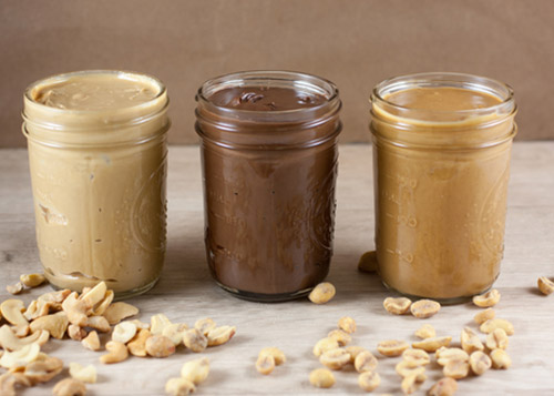 Three jars of different kinds of nut butter with peanuts strewn around on the table.