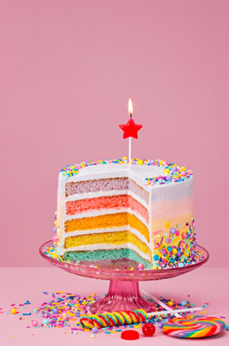 Rainbow themed-cake on a cake stand with candy and confetti below the stand