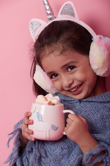 A girl wearing unicorn earmuffs holding a unicorn mug topped with marshmallows