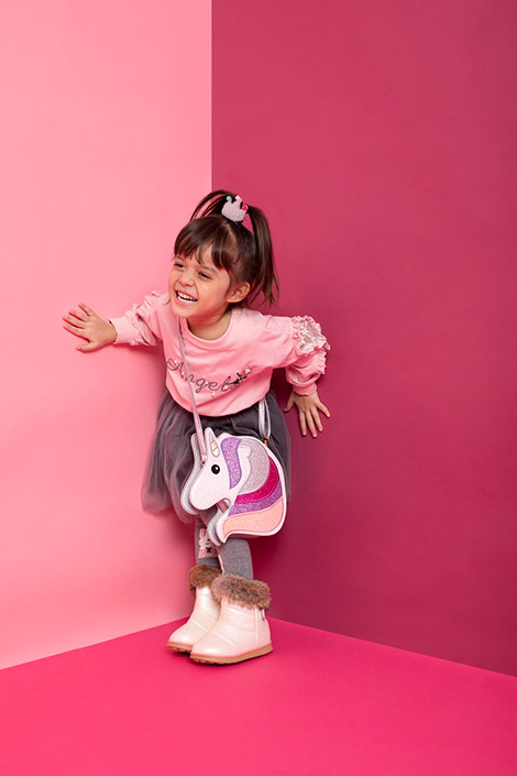  A girl standing against the wall and laughing while wearing a skirt, top, and a unicorn bag