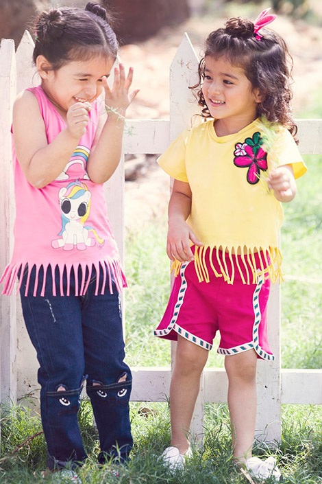 Two girls standing against a white picket fence and laughing. One of the girls is wearing a pink tee with a unicorn print