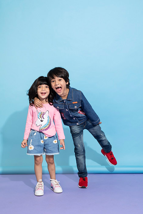 A girl and boy posing and smiling. The girl is wearing a pink sweatshirt with a unicorn print, the boy is wearing a denim jacket and denim jeans.