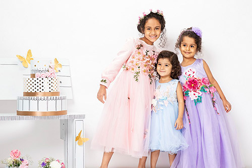 Three girls in pastel gowns standing next to a table with a cake