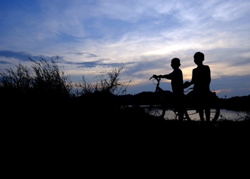 Two boys standing by a cycle in the dark.