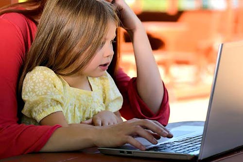 Daughter sitting on mom’s lap looking at the laptop on a table.
