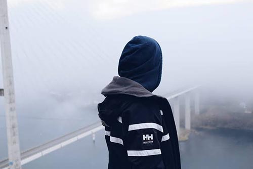 Child wearing a warm jacket with his back to the camera staring into the mist, looking out over a bridge in the winter.