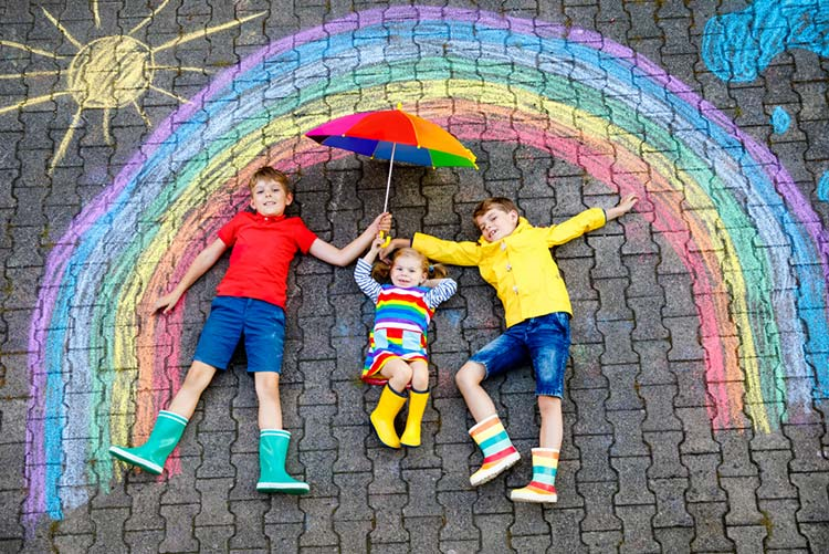 Two young boys and one toddler girl lying down on a cobbled street amidst a rainbow painted with crayons and an umbrella!