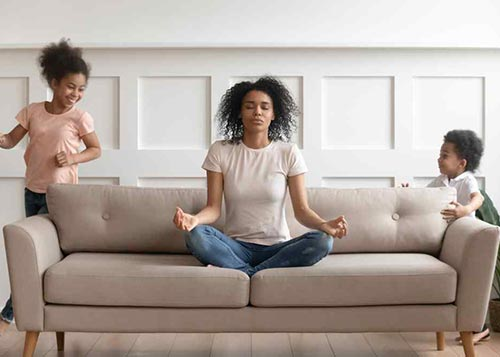 A mother meditating on the sofa with her two kids standing by the side smiling