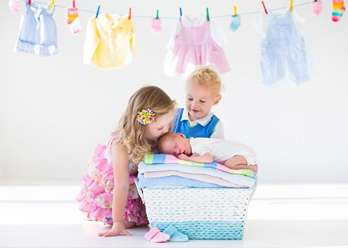 Two toddlers kissing an infant baby with kids’ clothes hanging in the background