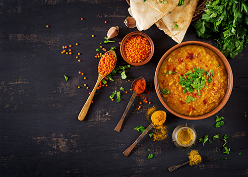 Bowl of lentil soup surrounded by fresh greens, Indian bread, and spoonfuls of Indian spices