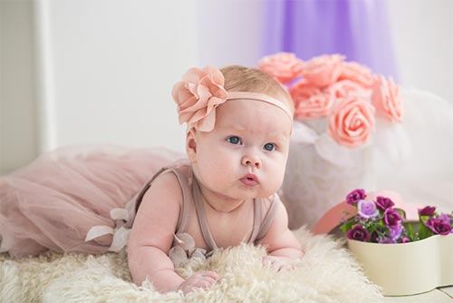 Adorable baby girl lying on a bed wearing a pink dress and pink flowers surrounded by purple roses