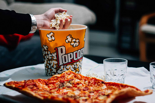 A hand grabbing popcorn from a popcorn bag with a pizza and glasses lying next to the popcorn bag