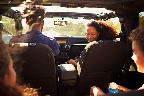 Parents and two kids in a car. Mother in the front seat is smiling at the kids in the backseat while dad drives. Sun roof is open, family is engaging in happy conversation.