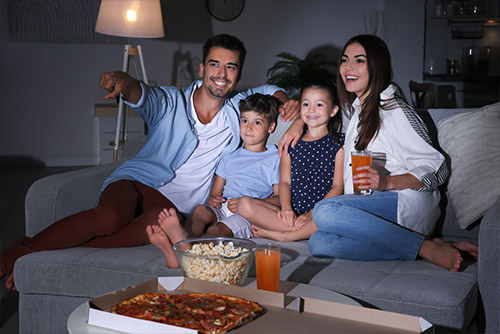 Mom, dad, son, and daughter sitting on the couch with a pizza, popcorn, and juice on the table. Dad is pointing at the TV (which not visible in the picture). 