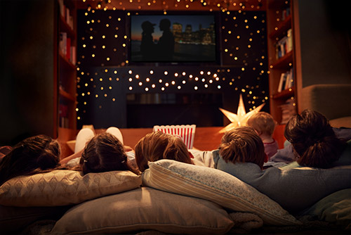 Mom and kids lying down next to each other watching a movie on the TV mounted to the wall with string lights around it