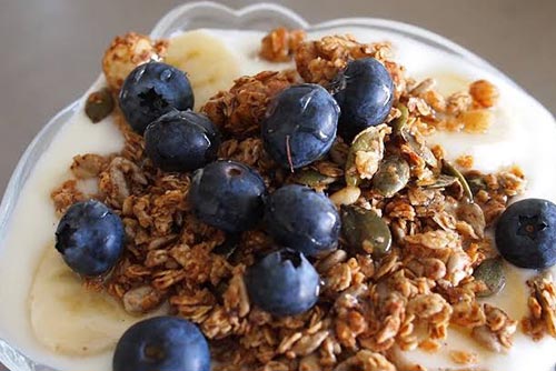 Full blueberries, cereal, sliced bananas, dry fruits like pista, almond, walnut, and sugar resting on a bed of fresh yoghurt.