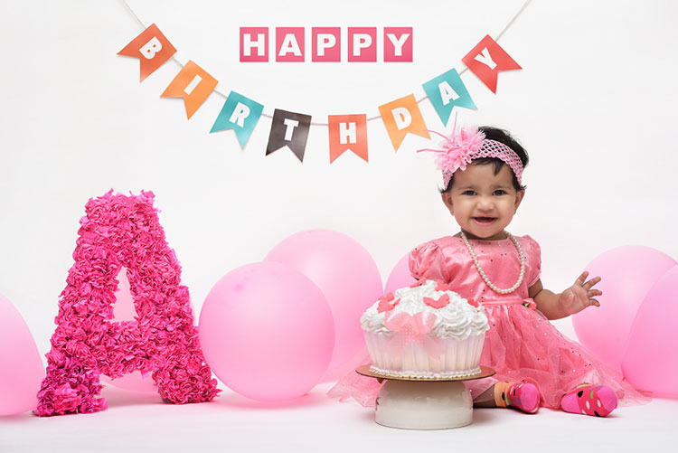 Birthday girl sitting with a cake, balloons, birthday streamers, and a rose standee of the letter A