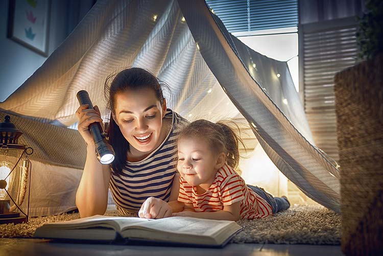 A mom and child under a lying down under a tent reading a story.