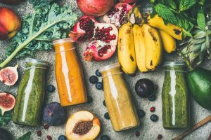 Smoothies in glass bottles surrounded by bananas, pomegranate, berries, apples, and greens