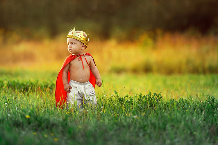 Adorable toddler wearing a king’s cape and crown, standing in the middle of a field!