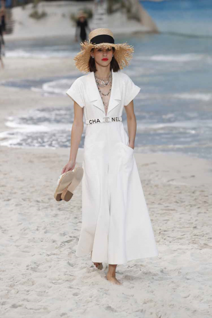 Woman walking on the beach wearing a white Chanel jumpsuit and beige and black hat, paired with accessories!
