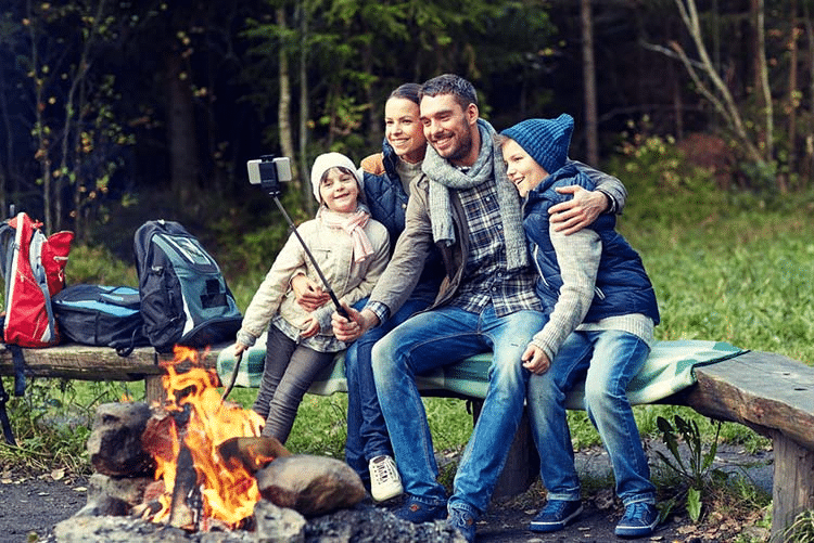Family sitting by the bonfire in the woods clicking a selfie with a selfie stick!