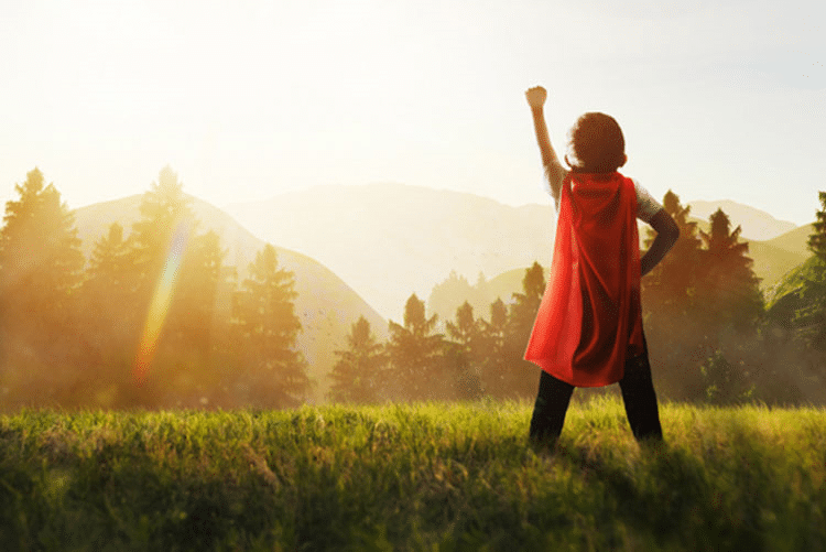 Child wearing a cape with his fist raised standing facing the mountains.