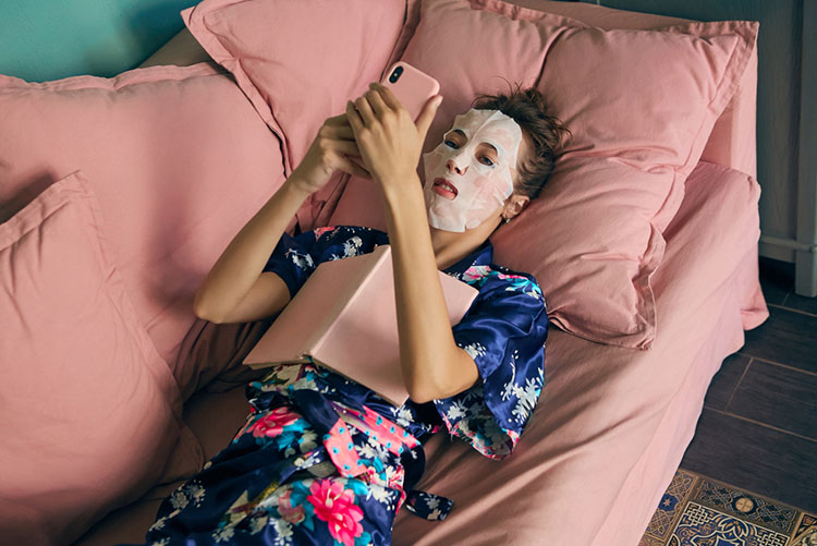 Woman wearing a sheet mask while lying down on the couch looking at her phone.