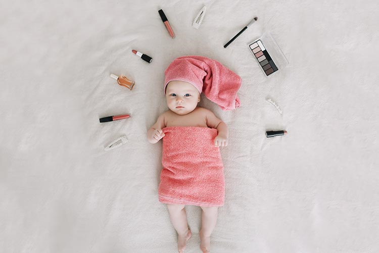 Infant wrapped in a towel, lying on a bed, surrounded by makeup objects such as lipstick, eyeshadow palette, makeup brush, etc.