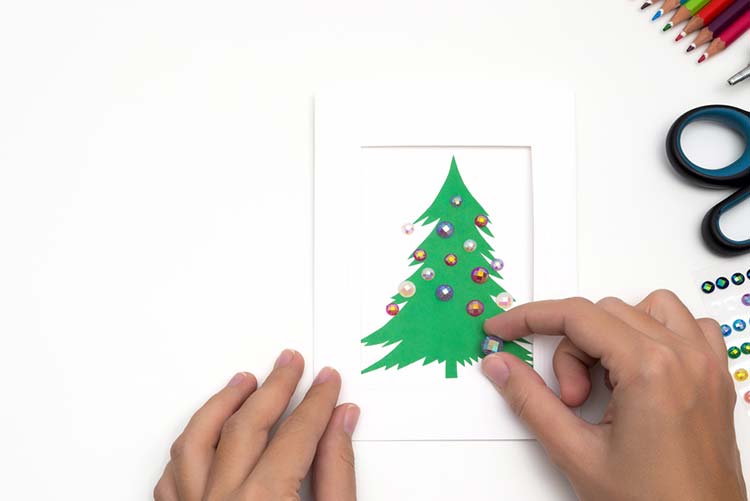Hands sticking a crystal button a Christmas tree on a card
