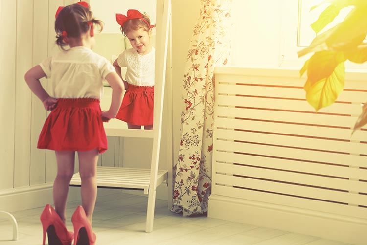 A little girl dressed up and standing in front of the mirror.