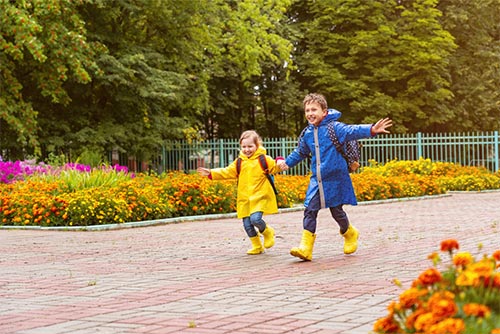 Two children dressed in raincoats and rain boots run while holding hands.