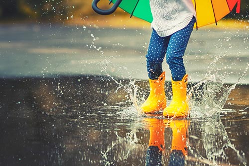 A child wearing rain boots jumps in a puddle of water.