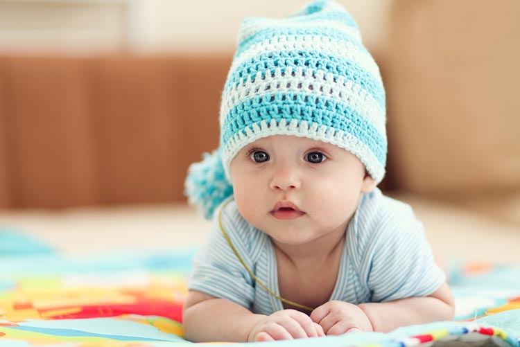 Adorable baby lying on a children’s rug wearing a white-blue woollen cap.