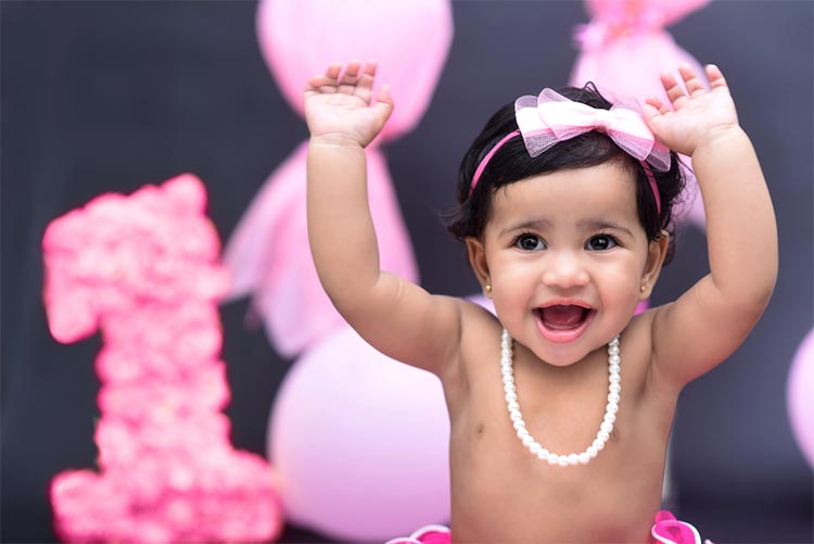 Adorable baby girl wearing a pink bow, skirt and a pearl necklace, throws her hands in the air while smiling at the camera.