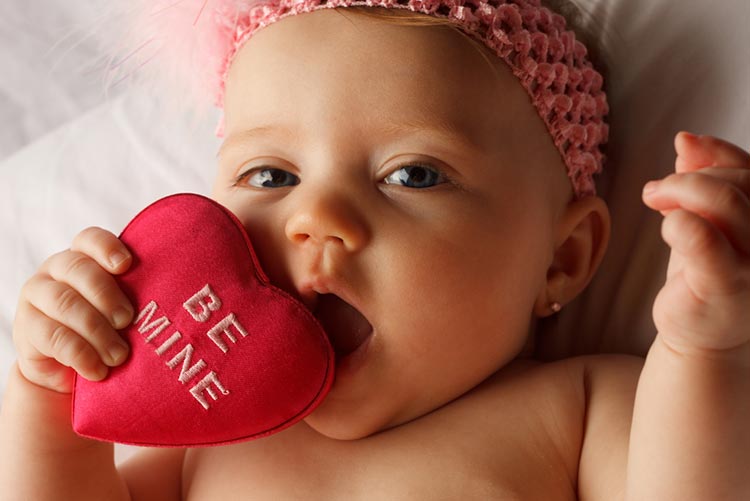 Baby girl holding a heart pillow that says ‘Be Mine’.