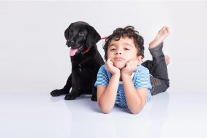 Adorable young boy sitting with hands on his cheeks alongside his black Labrador Retriever puppy!
