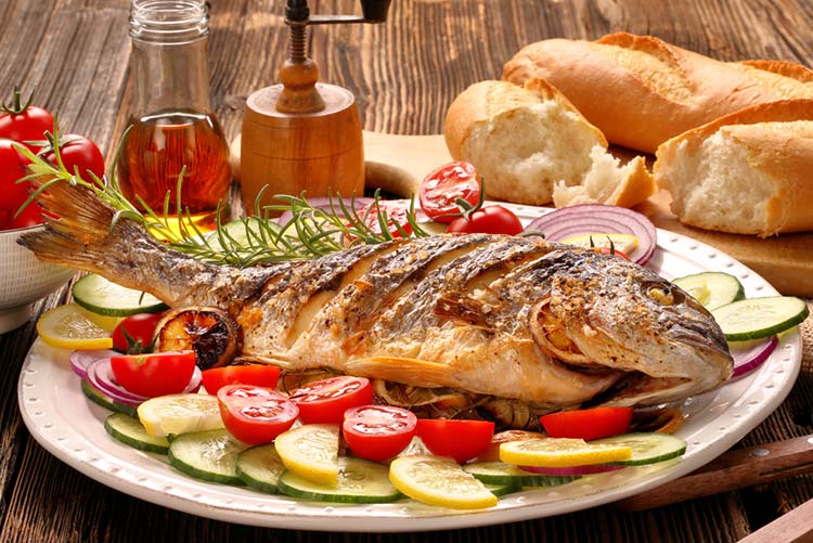 Whole cooked fish served on platter loaded with veggies such as tomatoes, onions, condiments, and baguettes.
